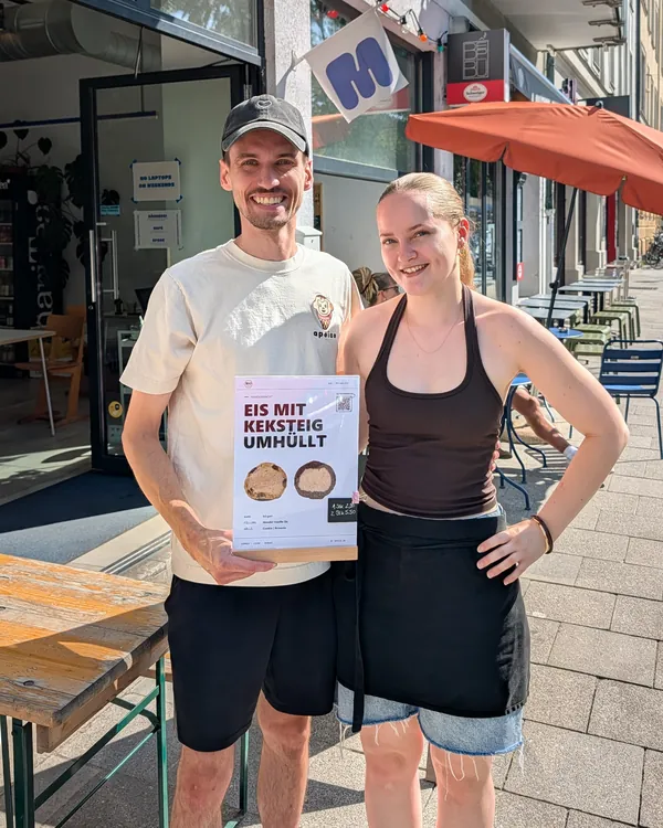 Joshua von apeice mit einer Mitarbeiterin von METTA und dem kü:gerl-Aufsteller vor dem Café